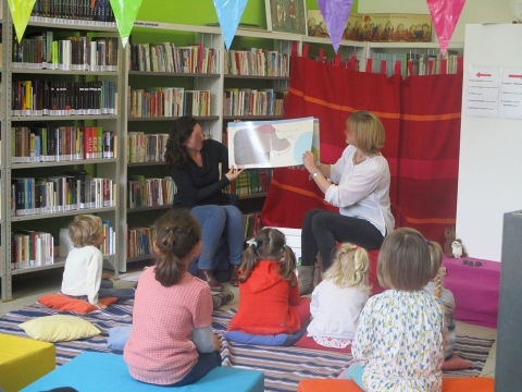 Photographie d'une bibliothécaire montrant un livre d'images à des enfants devant des rayonnages à l'intérieur de la bibliothèque de Sivry-Rance